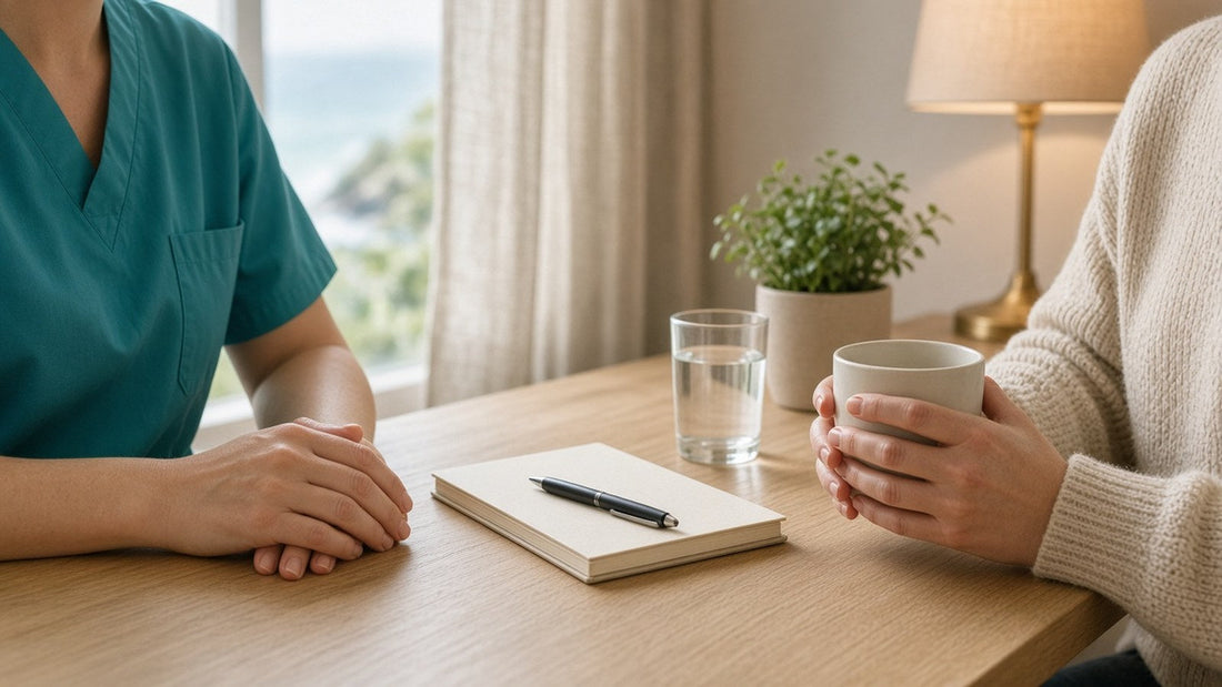 A nurse and a client talking at a light wood table with a notebook and a glass of water in a bright, calm consultation room